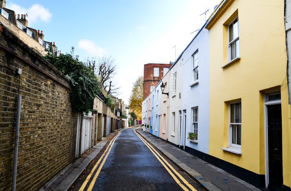 A residential street in Kentish Town lined with parked cars on both sides, including a mix of small hatchbacks and larger vans, with some vehicles featuring covers or storage containers. The pavement 