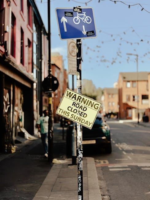 A street scene showing a metal pole with two signage boards: a blue bicycle and pedestrian path sign indicating a shared lane with an upward arrow for bicycles and a downward arrow for pedestrians, an