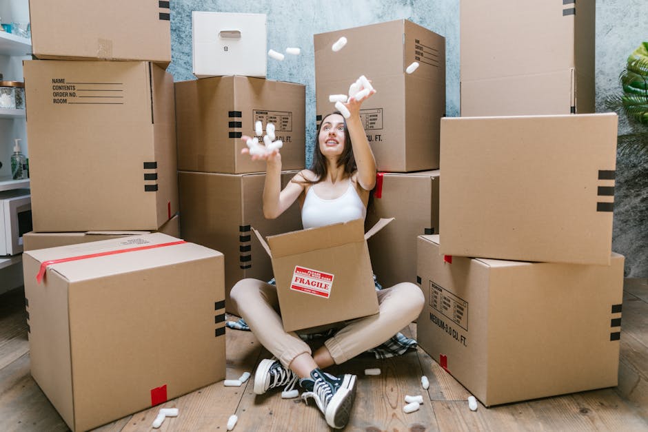 A young woman with dark hair and casual clothing is sitting cross-legged on a wooden floor inside a room, surrounded by numerous brown cardboard boxes of various sizes, some sealed with red tape and o