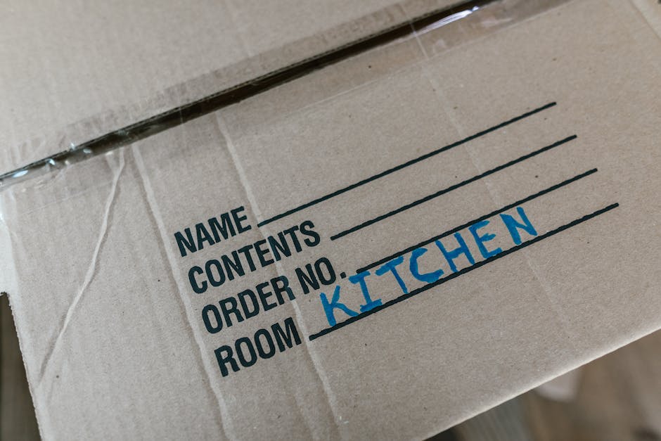 A young woman with dark hair and casual clothing is sitting cross-legged on a wooden floor inside a room, surrounded by numerous brown cardboard boxes of various sizes, some sealed with red tape and o